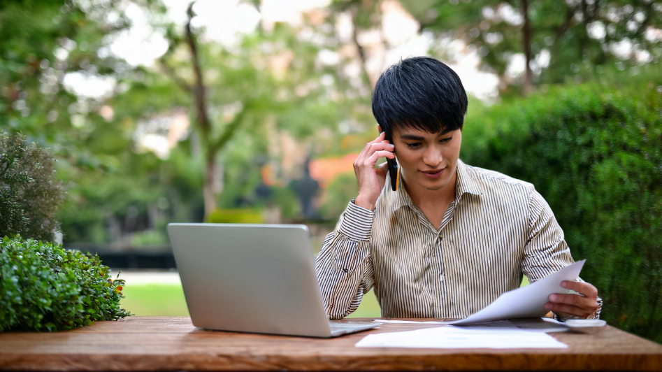 An entrepreneur working outdoors in a park, talking on the phone while reviewing documents with a laptop on the table, symbolizing the blend of work, creativity, and thoughtful planning in a serene environment.