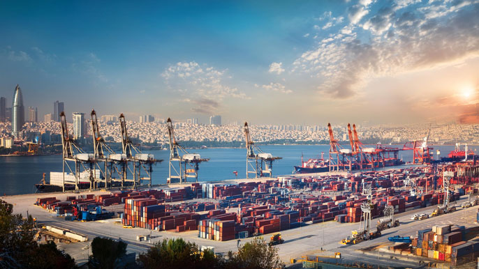 A busy trade port in Türkiye with cranes loading containers onto cargo ships, while trucks are parked nearby for transportation. The backdrop features a modern city skyline under a sky with scattered clouds, highlighting Türkiye's role in global trade.
